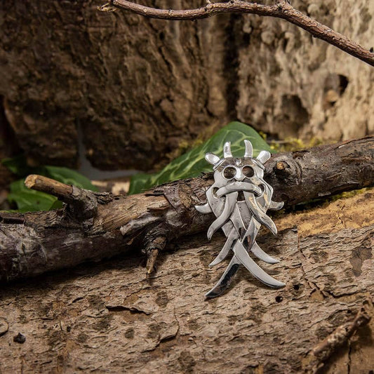 Et dramatisk Broche Maske 925s Sterling Sølv vedhæng med indviklede, flydende designs sidder på en ru, mosbevokset trægren i rustikke, naturlige omgivelser. Flere grønne blade og tekstureret bark tilføjer jordiske elementer til sammensætningen.