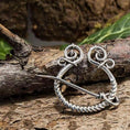 Indlæs billede i Gallery viewer, En rustik, håndlavet Broche Ringnål 925s Sterling Sølv med indviklede tråddesigns og cirkulære elementer hviler på en tekstureret trægren. Baggrunden har grønne blade og en ru, jordagtig overflade, der forstærker den naturlige æstetik.
