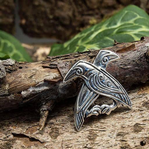 Broche Hugin og Munin 925s Sterling Sølv, formet som en sølvfugl og designet i keltisk stil, hviler på forvitret bark. Brochens indviklede detaljer, der minder om Odins ravne, forstærkes af barkens rustikke tekstur og blødt slørede grønne blade i baggrunden.