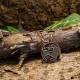 Indlæs billede i Gallery viewer, Wodan Mønt Vedhæng Bronze, et rundt og indviklet indgraveret vedhæng med design i vikingestil, hviler på en ru, barkbeklædt gren med grønne blade i baggrunden. Vedhænget udstråler en aura af antikken med sine gamle, mørklagte bronzedetaljer.
