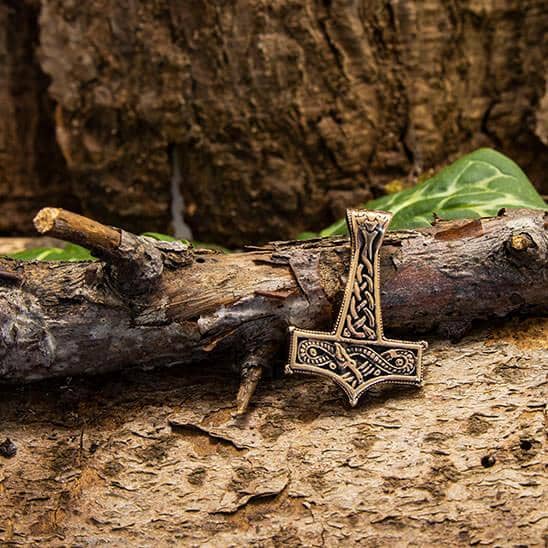 Et detaljeret, rustikt vedhæng formet som Thors Hammer hviler på en forvitret trægren. Baggrunden har struktureret brun bark og spredte grønne blade. Thors Hammer Midgårdsormen Vedhæng Bronze vedhæng har indviklede, symmetriske designs og en aura, der minder om Midgårdsormen, hvilket tilføjer dens mystiske tiltrækningskraft.