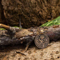 Indlæs billede i Gallery viewer, En rund amulet Frigg Vedhæng bronze med indviklede graveringer hviler på en struktureret træoverflade, på en baggrund af træbark og grønne blade. Designet på vedhænget har detaljerede og symmetriske mønstre, hvilket tilføjer dens rustikke charme.
