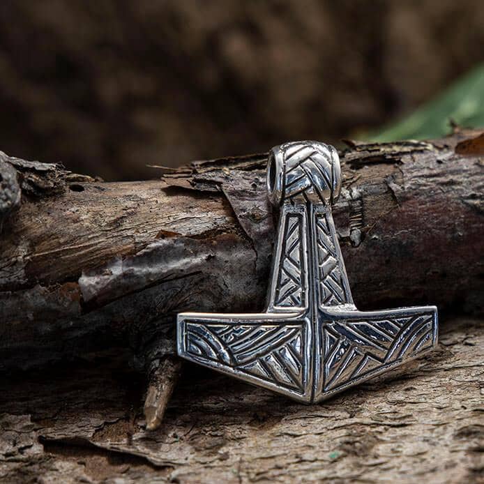 Thors Hammer Vedhæng Sindri 925s Sølv, et sølvvedhæng, der forestiller en stiliseret Mjölnir (Thors Hammer) med indviklede graveringer, hviler mod en struktureret træbarkbaggrund. Vedhænget er lavet af 925s Sterling Sølv og har detaljerede symmetriske mønstre, der fremkalder nordiske mytologi.