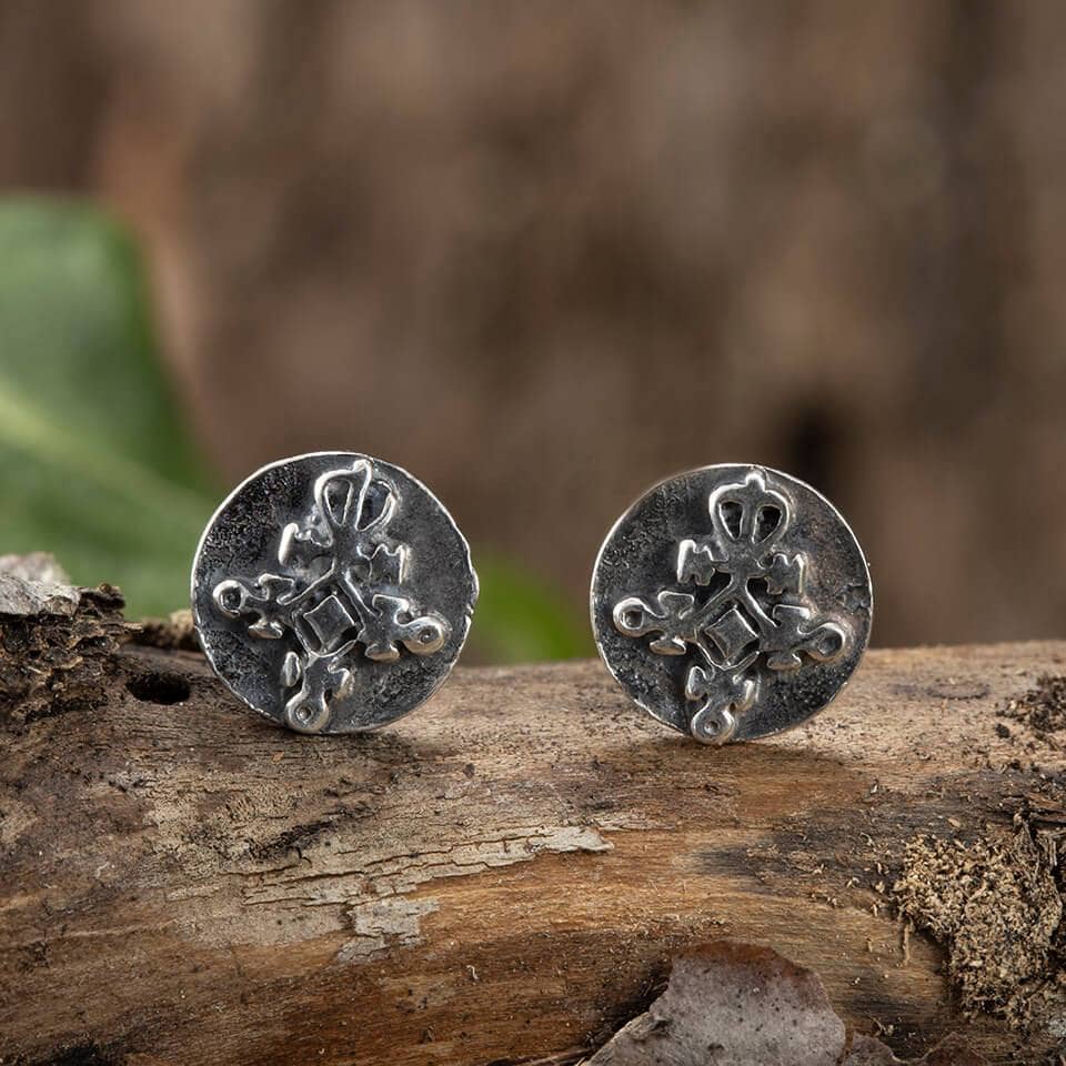 Et par Amulet Love Studs P 925s Sterling Sølv manchetknapper med indviklede krydsdesigns i middelalderstil vist på et stykke rustikt træ med en sløret grøn baggrund.