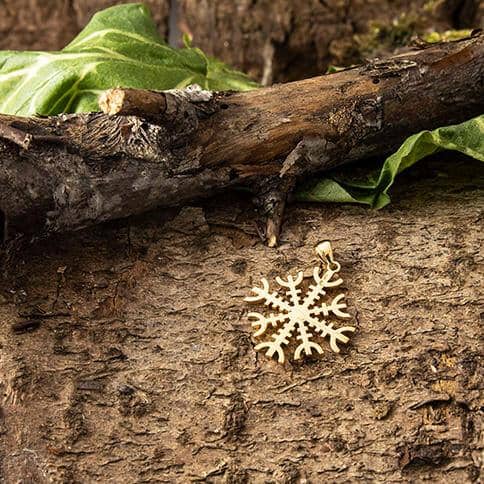 En Rune Amulet Ægishjalmur Vedhæng Forgyldt 925s Sterling Sølv ligger på en ru, brun træoverflade, delvist dækket af en struktureret nedfalden gren og grønne blade i baggrunden.