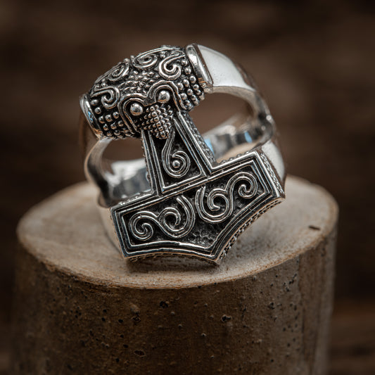 En Sølvring Thors Hammer Mjølnir 925s Sølv i ægte Sterlingsølv med et detaljeret Thors hammerdesign med indviklede nordiske mønstre hviler på et cylindrisk træstativ. Hammerens hoved er prydet med hvirvelmotiver og perledetaljer, der understreger dets detaljerede mønster og håndværk.