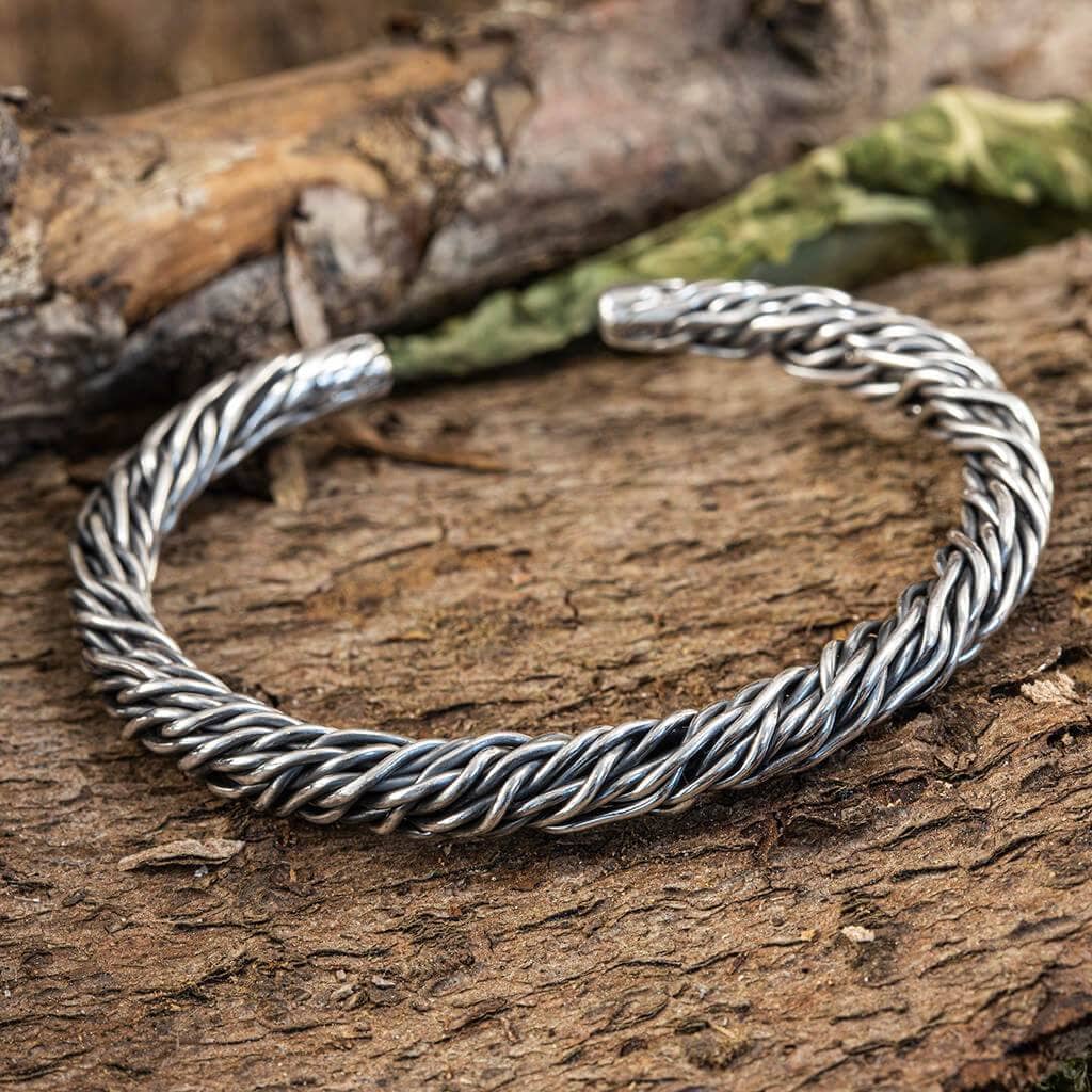 Et Armbånd Bangle Mesh 925s Sterling Sølv er placeret på en tekstureret træoverflade. Armbåndet har et indviklet flettet design, der fremkalder en rustik og jordnær æstetik. Baggrunden inkluderer antydninger af grønt og bark, hvilket forstærker det naturlige tema.