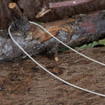 Indlæs billede i Gallery viewer, Halskæde Slangekæde 925s Sølv 1mm er draperet over en ru trægren, der ligger på en skovbund. Baggrunden inkluderer bark, snavs og nedfaldne blade, hvilket bidrager til en naturlig, robust indstilling.
