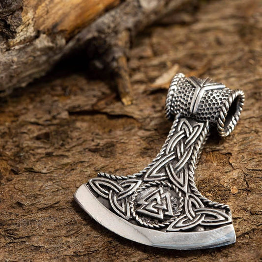 En detaljeret Thors Hammer Vedhæng Økse lavet af 925'erne Sterling Sølv, med indviklede keltiske og vikinge-inspirerede designs med knudemønstre og geometriske symboler, hviler på en struktureret, stenet overflade. Vikingesmykke fremviser enestående håndværk og kunst på baggrund af ru natursten.