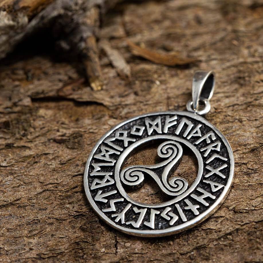 Et cirkulært sølv Runeamulet Vedhæng 925s Sølv vedhæng prydet med runeindskrifter i gammelt vikingealfabeter rundt om kanten og et smart tredobbelt spiralsymbol i midten, hviler på en struktureret træoverflade. Vedhænget er lavet af Sterling Sølv og har en løkke øverst til fastgørelse til en kæde.