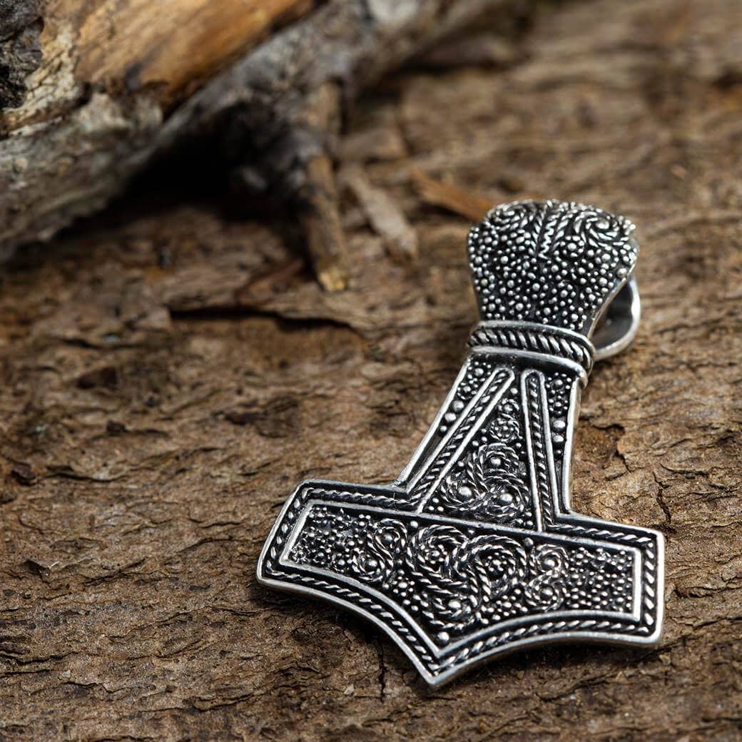 En detaljeret, sølvfarvet Thors Hammer Vedhæng Borrestil 925s Sterling Sølv hviler på en struktureret, jordagtig træoverflade. Det indviklede design inkluderer udsmykkede mønstre og prikker, der fremhæver dets håndværk. Dette vedhæng er et vidnesbyrd om udsøgt kunst.