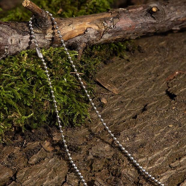 En skinnende, nikkelfri Halskæde Kuglekæde Stål ligger på jorden blandt pletter af grønt mos og brun, struktureret træbark. Den enkle kæde står i kontrast til de naturlige, jordiske elementer, der omgiver den.