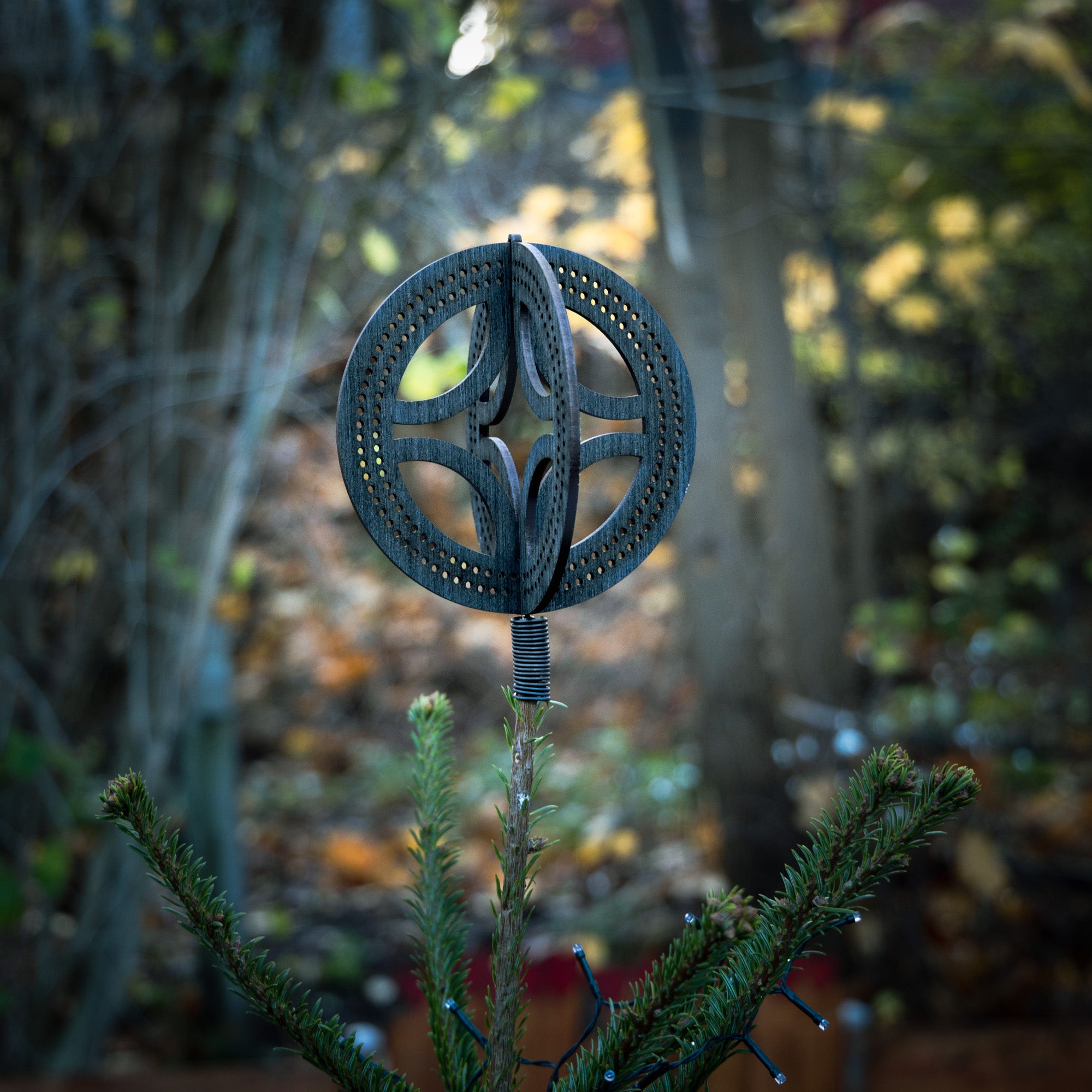 Et nærbillede af den karakteristiske Solhjul Trætop / Julestjerne (18cm), der genlyder designs fra oldnordisk kultur, elegant pryder et stedsegrønt træ. Den slørede baggrund viser en skov med efterårsblade, der forstærker den fredfyldte og naturlige atmosfære.