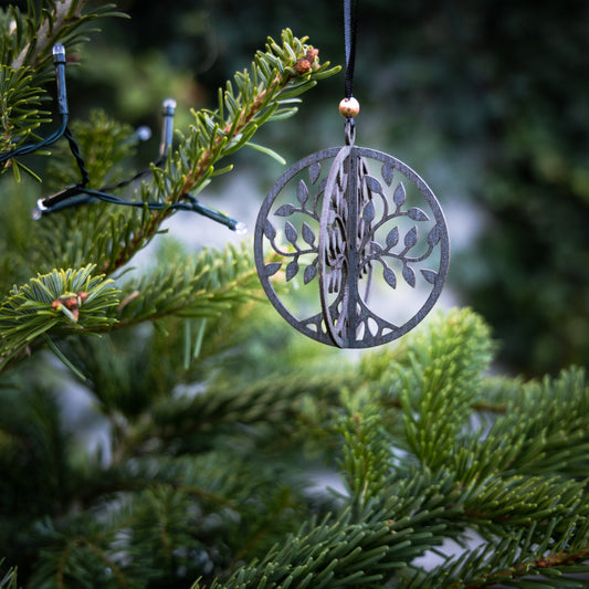 Et Mobil Yggdrasil Wood Viking metal ornament, der forestiller det mytiske træ fra nordisk mytologi med grene og blade, hænger fra en sort snor på et grønt fyrretræ. Fyrretræets grene er prydet med uoplyste snorelys. Baggrunden er sløret og fremhæver ornamentet og fyrrenålene.