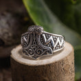 Indlæs billede i Gallery viewer, En detaljeret Sølvring Thors Hammer 925s Sølv, der viser et indviklet vikinge-inspireret design, sidder på en træoverflade. 925s Sterling sølv-ringen har udsmykkede mønstre, der ligner Thors hammer og er omgivet af grønt i den slørede baggrund.
