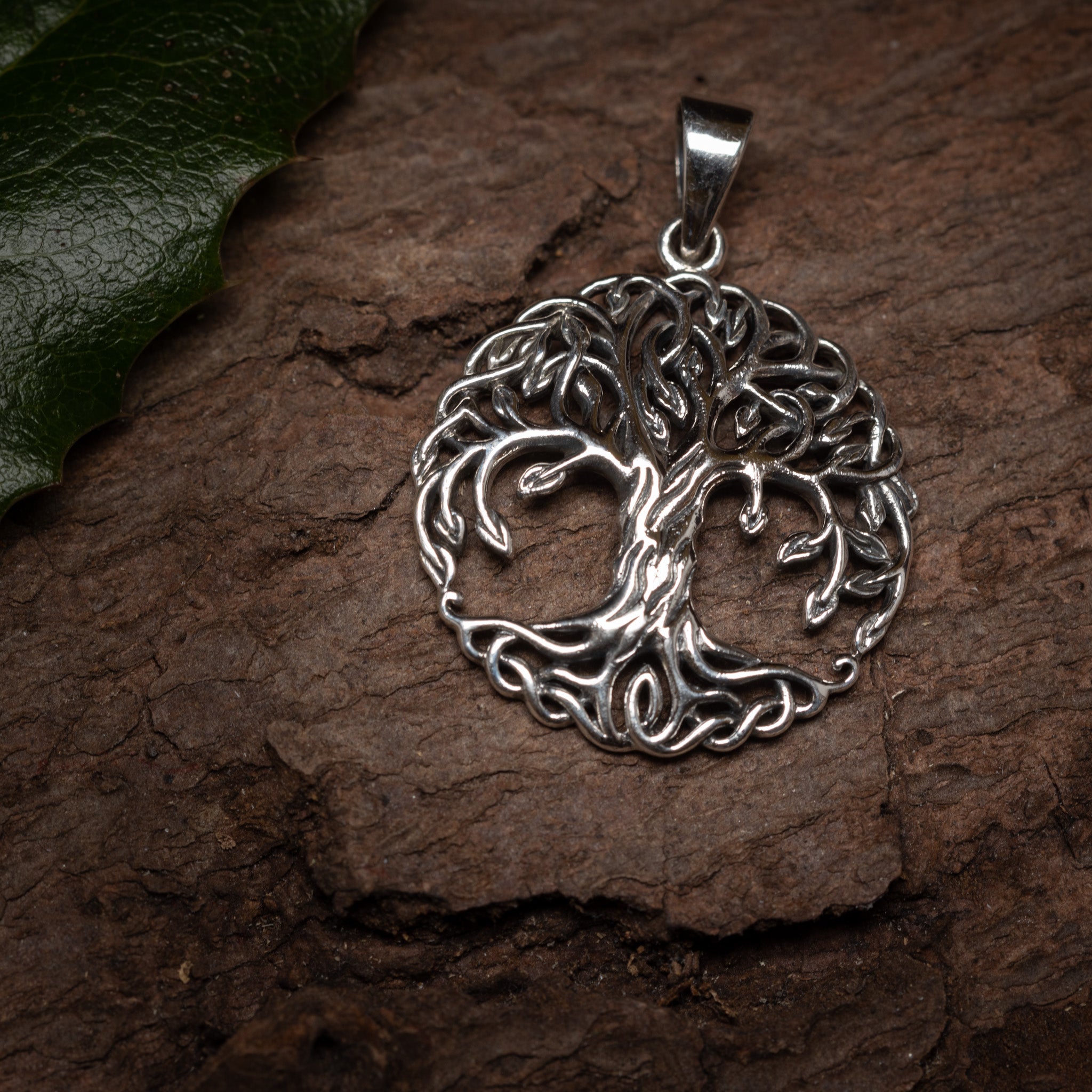 Yggdrasil Livets Træ Tüngl Vedhæng 925s Sterling Sølv vedhæng med et detaljeret Tree of Life-design ligger på en tekstureret træoverflade. Den cirkulære vedhæng viser indviklet snoede rødder og grene. Et grønt kristtornblad er synligt i øverste venstre hjørne af billedet, hvilket påkalder et strejf af nordiske mytologi.