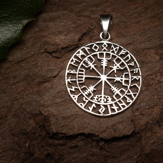 Vedhæng Vejviser m. Runer 925s Sølv vedhæng, med indviklede Furthark alfabetsymboler og et centralt Vegvisir kompasdesign, ligger på en tekstureret træoverflade. Et grønt kristtornblad er delvist synligt i øverste venstre hjørne.