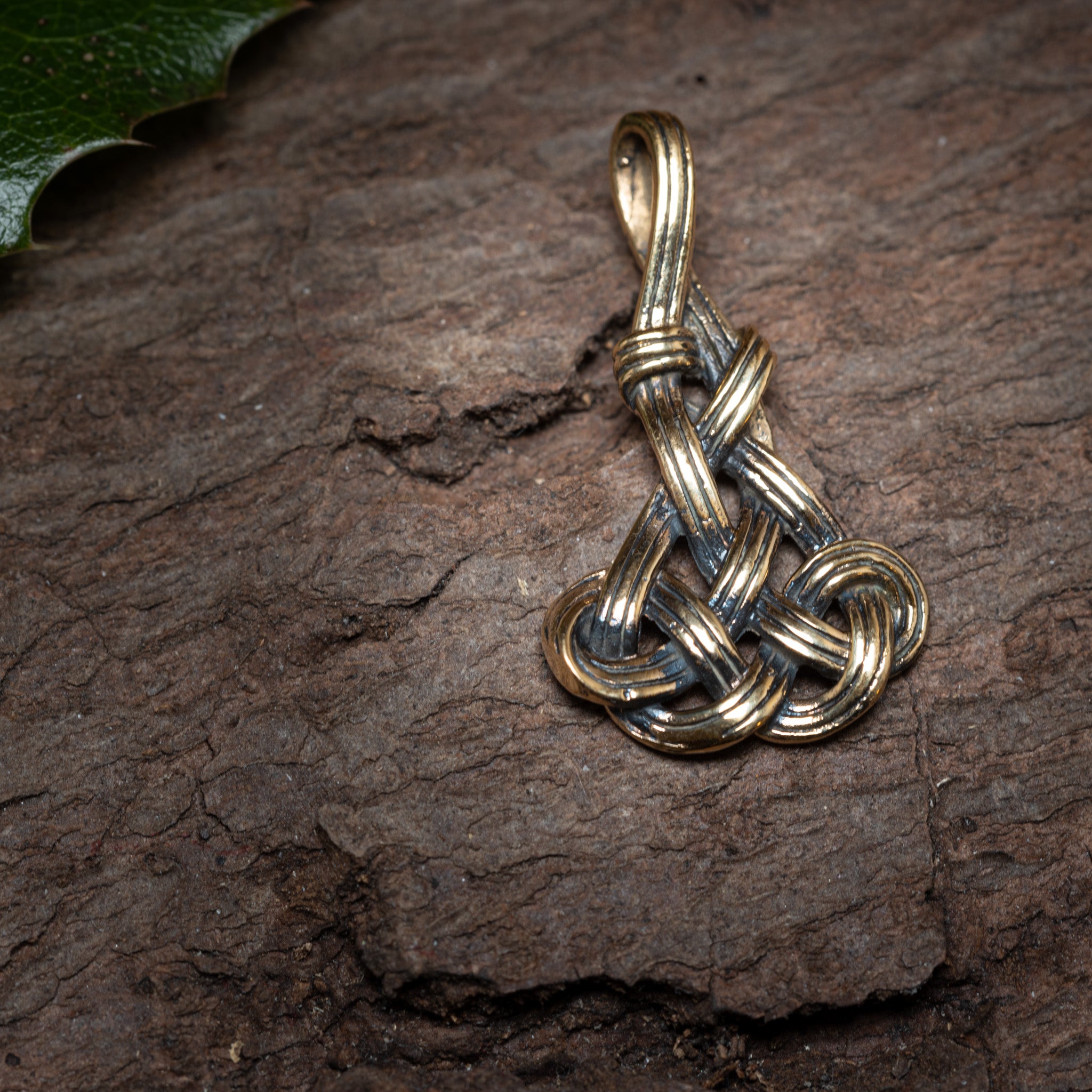 En detaljeret Keltisk Knude Vedhæng Knot Bronze i form af et nordisk knudesymbol hviler på en tekstureret træoverflade. Til venstre er en del af et grønt blad synligt, hvilket tilføjer et naturligt element til sammensætningen. Det indviklede design fremhæver elementer, der minder om traditionelle keltiske smykker.