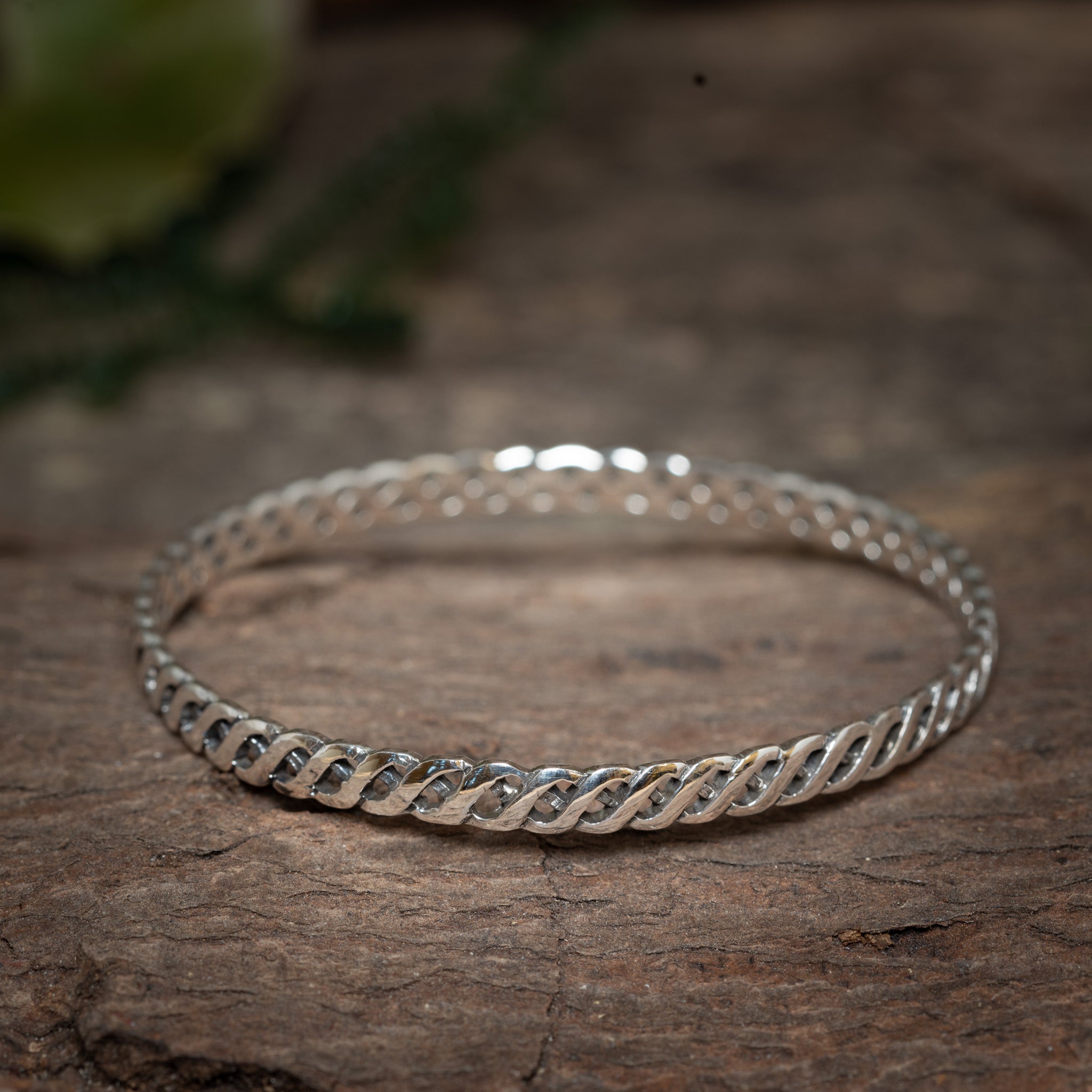 Et nærbillede af den delikate Armbånd Bangle Löft 925s Sterling Sølv med et indviklet flettet design hvilende på en struktureret træoverflade. Det nordiske vævet mønster skiller sig smukt ud, og baggrunden er let sløret, hvilket gør opmærksom på armbåndets detaljerede håndværk.