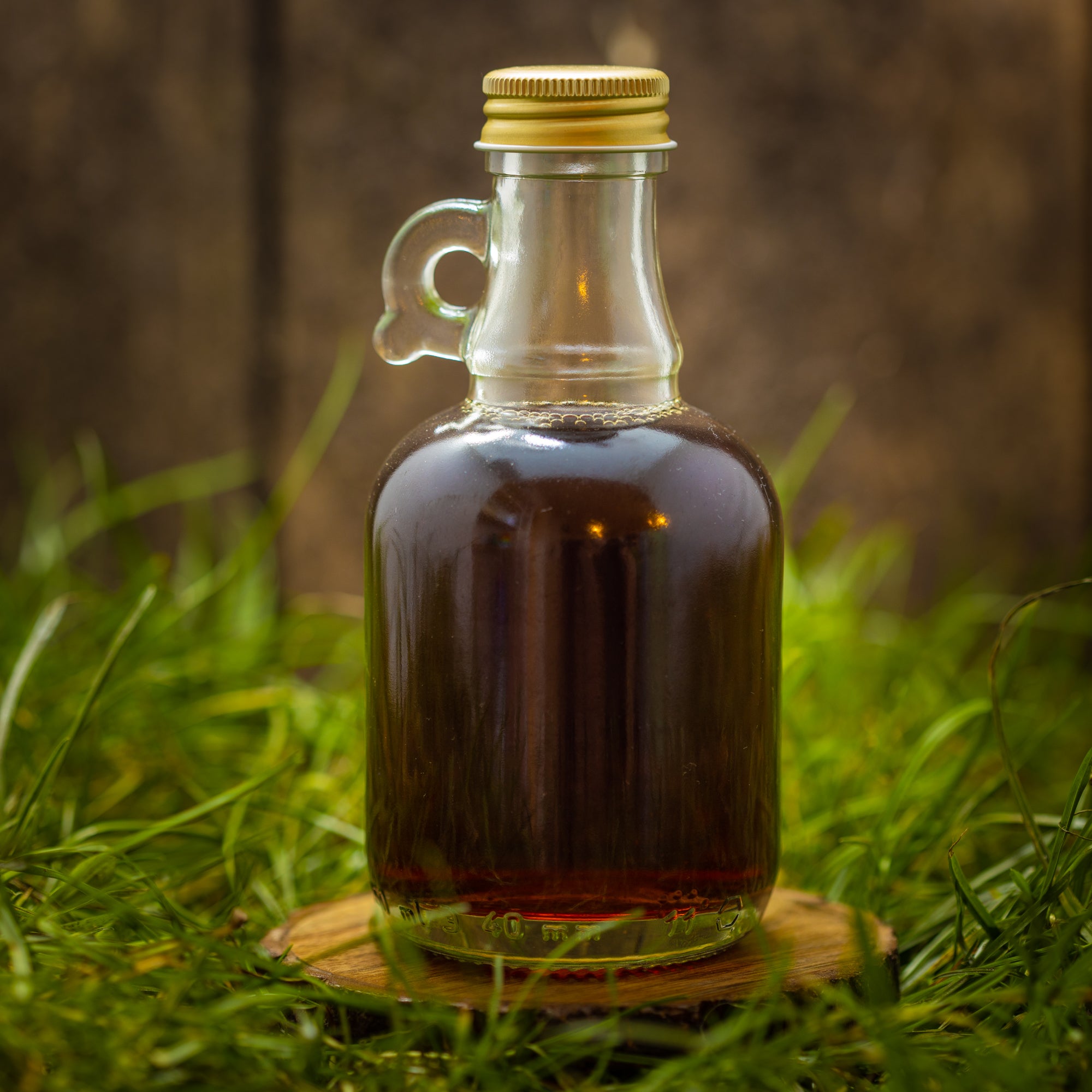 En lille, klar glasflaske med en gylden hætte rummer en mørk væske, der ligner ahornsirup. Flasken, mærket Dansk Mjød Jyde Mjød (200 ml), står på en træskranke midt i grønt græs, med en sløret, naturlig baggrund.