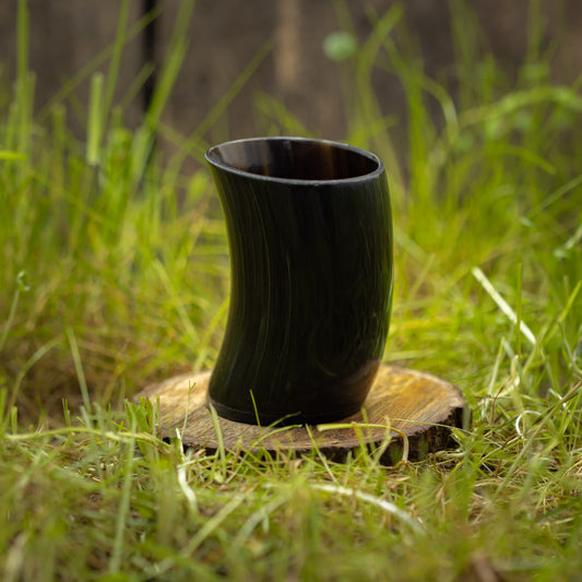 Et mørkt, buet Drikkeglas Nature Horn (100ml) står oprejst på en rund træbund, placeret på grønt græs. Baggrunden er sløret, men antyder en naturlig udendørs indstilling.