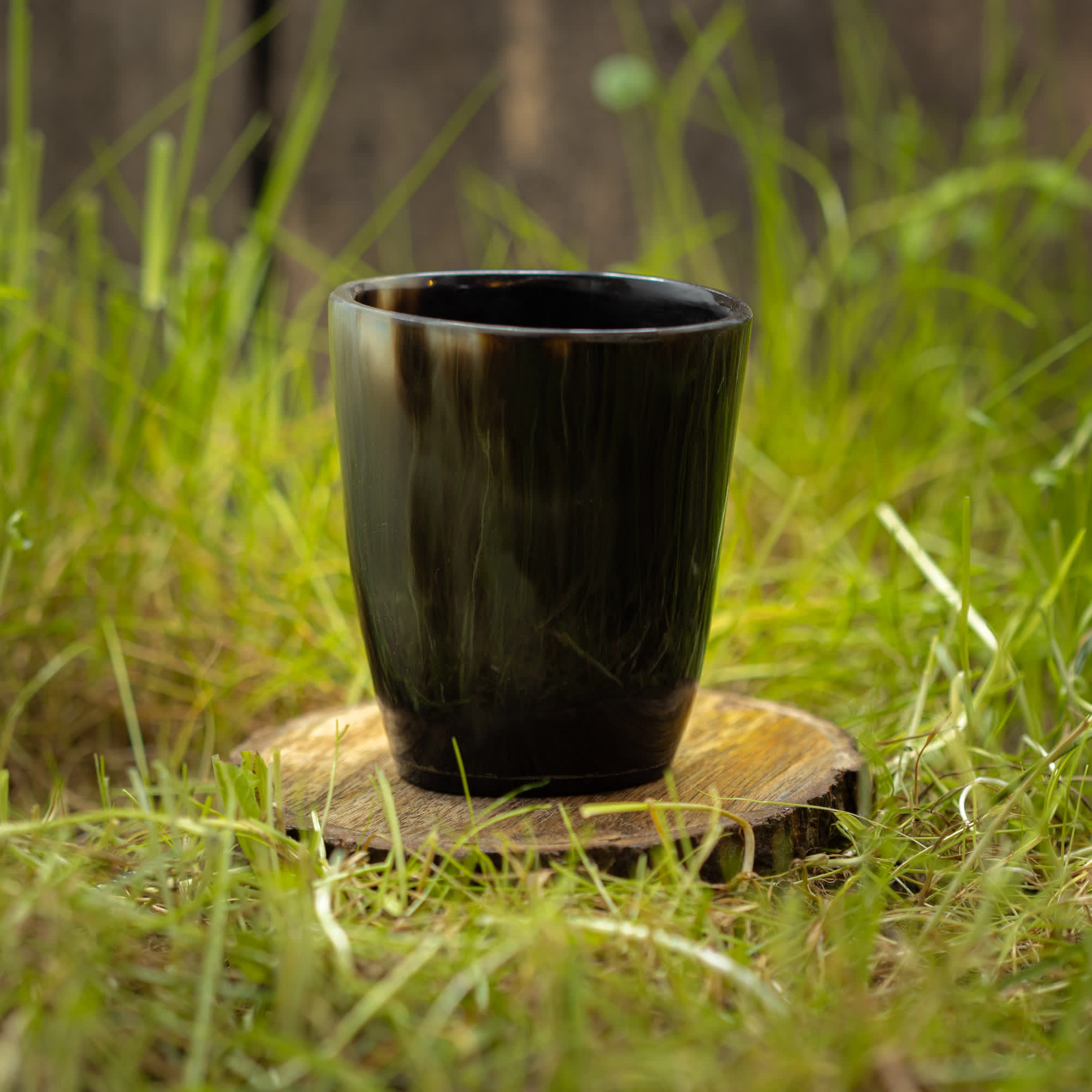 En mørk keramisk kop sidder på en træskranke midt på en græsmark. I den lidt slørede baggrund tilføjer Drikkeglas Nature Horn (100ml) et ekstra strejf af rustik charme, fremhæver de naturlige omgivelser og skaber en rolig atmosfære.