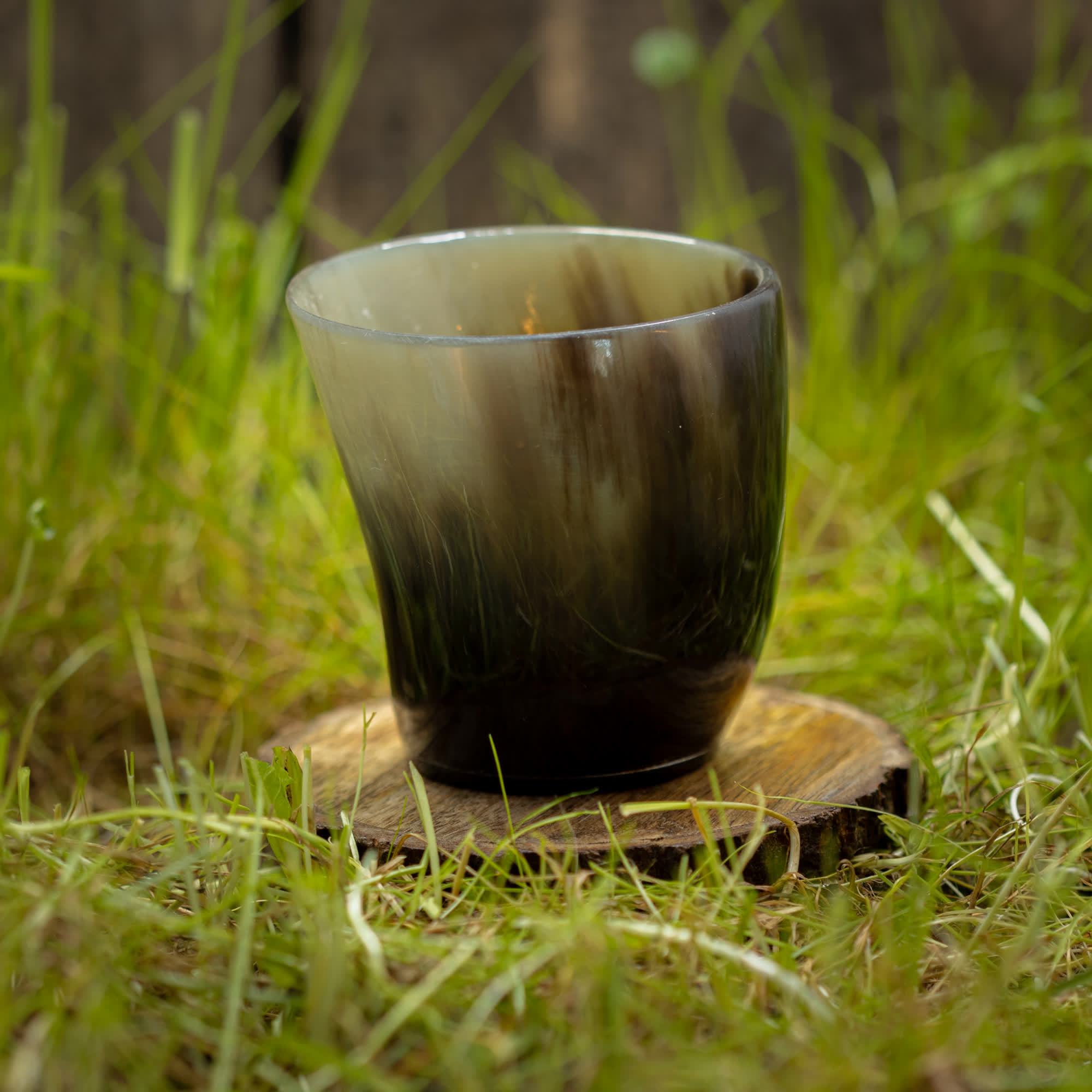 Et Drikkeglas Nature Horn (100 ml) sidder på en træskranke, omgivet af græs. Koppen har en naturlig gradient fra lys til mørk, hvilket fremhæver dens unikke tekstur og farvevariationer. Billedet er taget udendørs, i rustikke og naturlige omgivelser.