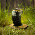 Indlæs billede i Gallery viewer, Et udskåret Drikkeglas Horn med Mønster (100ml) med en guldkantet kant sidder på en træskive midt i frodigt grønt græs. Drikkehornet fremviser indviklede geometriske mønstre og kan prale af en poleret, skinnende overflade. Baggrunden byder på sløret grønt græs og et rustikt plankeværk.
