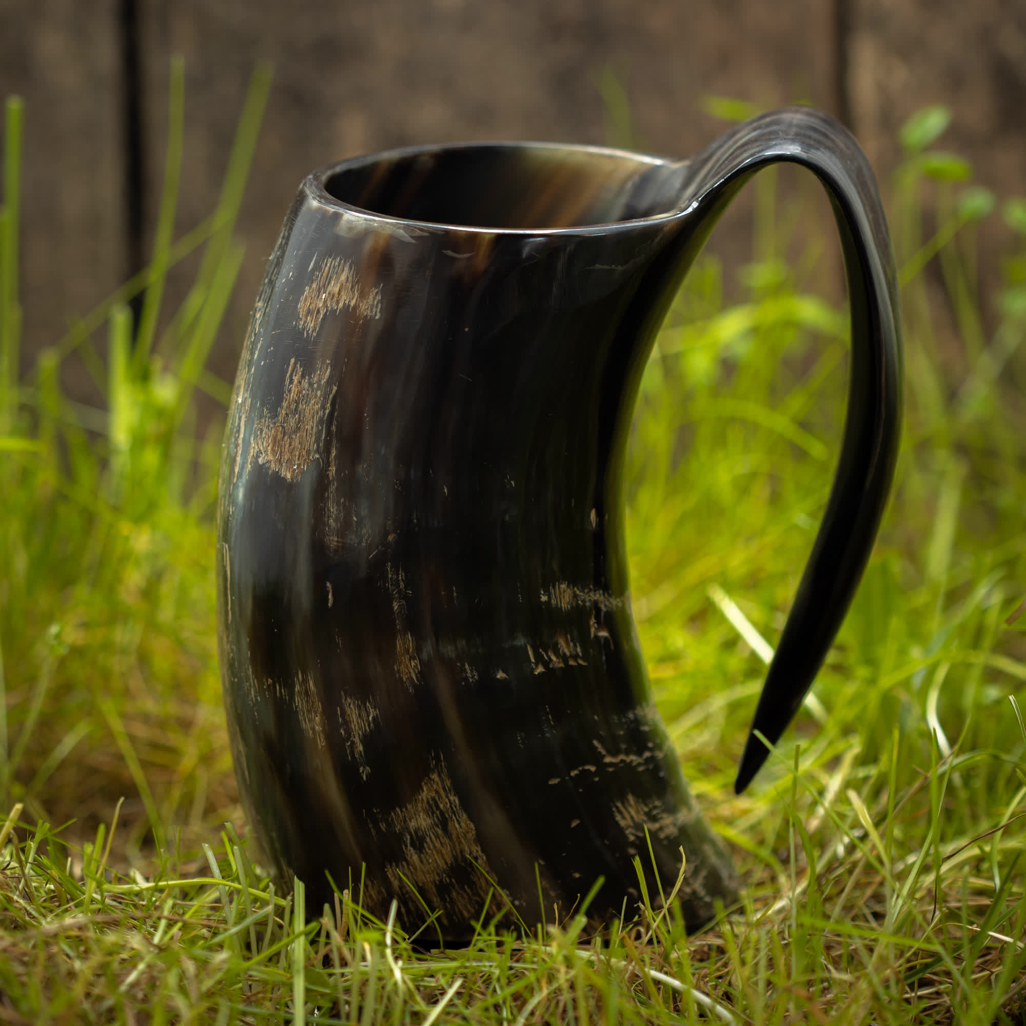 Et Vikingekrus Nature Horn (600-800ml) med et buet håndtag sidder på græs. Det håndlavede, polerede horn har en blank, mørk overflade med lysere pletter, der fremviser en naturlig og rustik æstetik. Baggrunden, der minder om vikingekrus, er let sløret for at understrege hornets detaljerede tekstur og form.