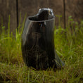 Indlæs billede i Gallery viewer, Et Vikingekrus Thors Hammer Horn (600-800 ml) med indviklede udskæringer, der ligner Thors hammer, sidder på grønt græs. Dette sorte drikkehorn står stolt på baggrund af forvitrede træplanker og tilføjer et rustikt præg til scenen.
