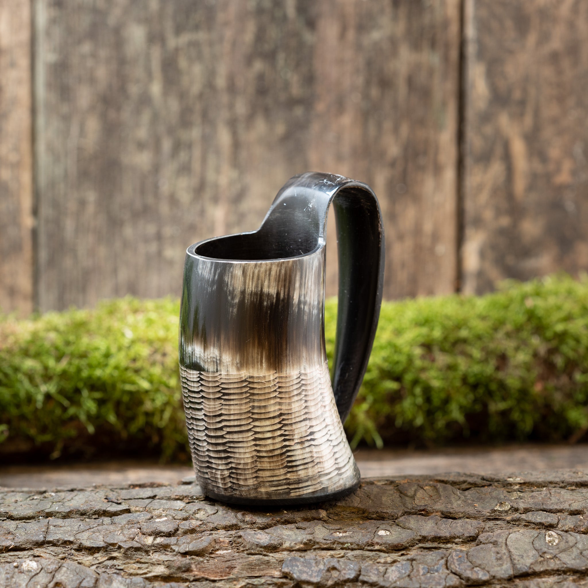 En hornformet Mini Vikingekrus Horn Rifflet (100ml) med et håndtag, hvilende på en rustik træoverflade. Karret har en struktureret base og en glat overdel med gradueringer fra sort til hvid. Baggrunden er sløret, med et plankeværk og grønt mos, der minder om Roskilde showrooms autentiske stemning.