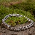 Indlæs billede i Gallery viewer, Et Armbånd ByKila Grov Rumpeflet 7mm 925s Sølv, komplet med sin indviklede lås, er elegant udstillet på en overflade af grønt mos og stykker af bark. Denne indstilling forstærker den naturlige og jordiske følelse af det polerede, flettet mønster smykke.
