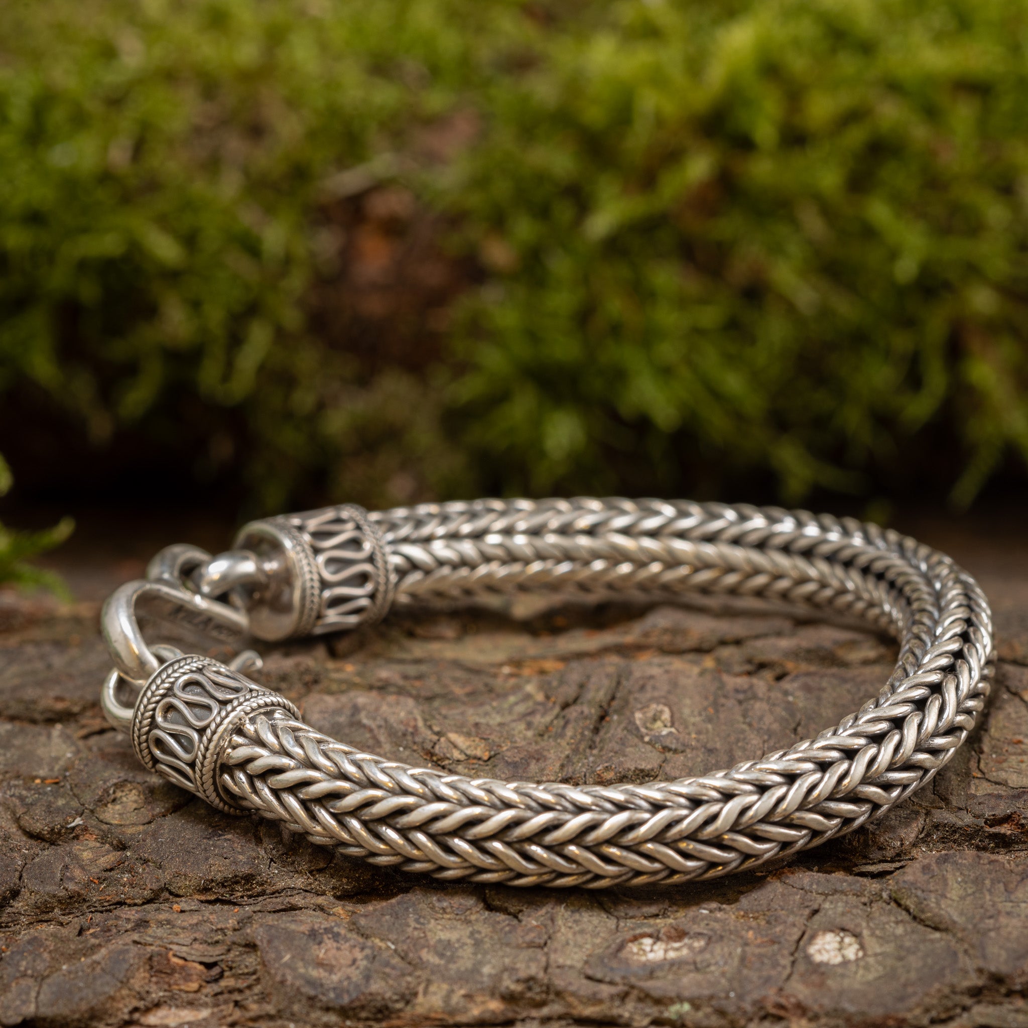 Armbånd ByKila Sildeflet 8mm 925s Sølv, med indviklede mønstre og lavet af 925s Sterling Sølv, er elegant placeret på en tekstureret træoverflade. Armbåndet viser udsmykkede endestykker og en klassisk kroglås på baggrund af frodigt grønt mossyet løv.