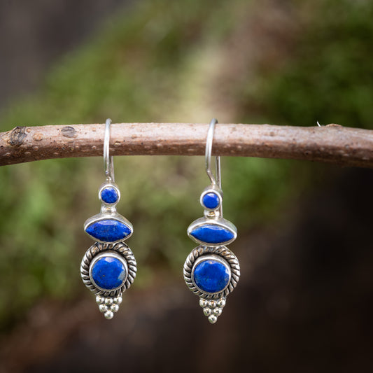 Et par Móðir Lapis Lazuli Hængeøreringe lavet af 925'er Sterling Sølv, med poleret lapis lazuli sten og indviklede designs, hænger fra en slank trægren mod en sløret, grøn naturlig baggrund.