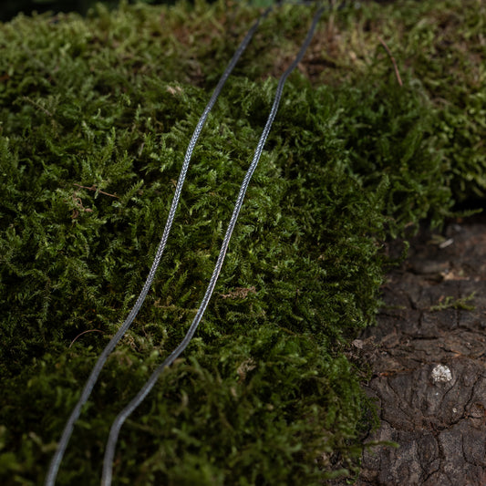 Nærbillede af en Halskæde Rævehale 925s Sølv 1,6mm liggende på en bund af grønt mos og et stykke bark i naturlige udendørs omgivelser. Tråden, der ligner middelaldersmykker, fremstår forvitret og står i kontrast til det levende mos.