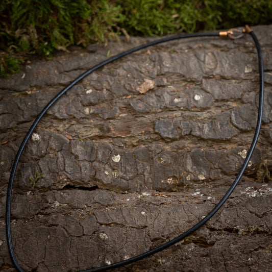 Et slankt sort kabel med et lille stik i den ene ende er placeret mod en struktureret træbarkoverflade, hvilket fremkalder den robuste skønhed, der minder om Læderhalskæden Tyk Glat Bronze. Baggrunden har ude af fokus grønt, måske mos eller løv.