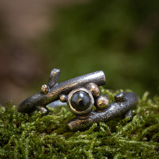 "Dark Beauty"-ringen af Eryn Design er sat mod en grøn mosbevokset baggrund. Denne fingerring i sterling sølv har et kvistlignende bånd prydet med små gyldne og sølvperleaccenter, der omkranser en central mørk hæmatit ædelsten. Det organiske design blander sig harmonisk med dets naturlige omgivelser.