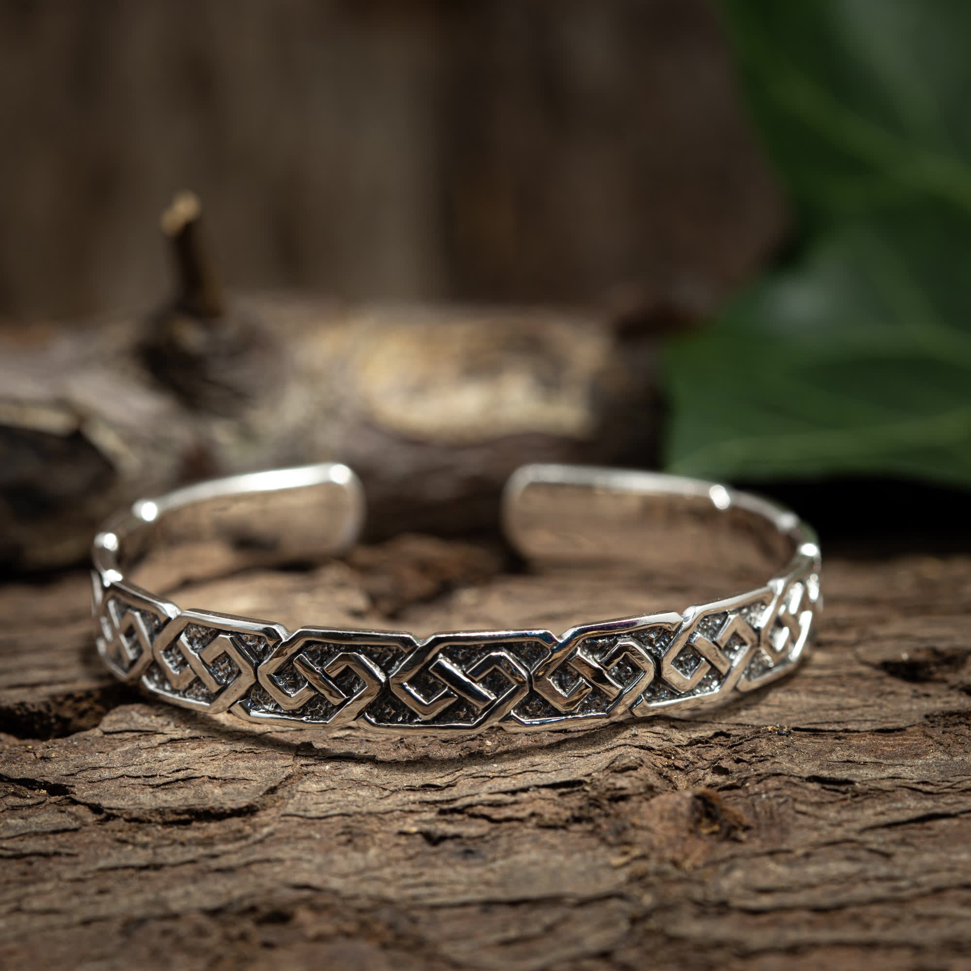 Et Armbånd Bangle Chief 925s Sterling Sølv-armbånd med et indviklet, sammenvævet keltisk knudedesign vises på en rustik træoverflade. Et sløret stykke træ og grønt løv er i baggrunden, hvilket fremhæver vikingetidens mønster på dette justerbart armbånd.