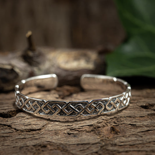 Et Armbånd Bangle Chief 925s Sterling Sølv-armbånd med et indviklet, sammenvævet keltisk knudedesign vises på en rustik træoverflade. Et sløret stykke træ og grønt løv er i baggrunden, hvilket fremhæver vikingetidens mønster på dette justerbart armbånd.