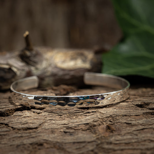 Armbånd Bangle Vlyi 925s Sterling Sølv, et sølv, tekstureret armbånd, er vist hvilende på en rustik træoverflade. Baggrunden har en sløret visning af en log og grønne blade. Dette åbent armbånd har et hamret design, hvilket giver det et unikt og håndværksmæssigt udseende.