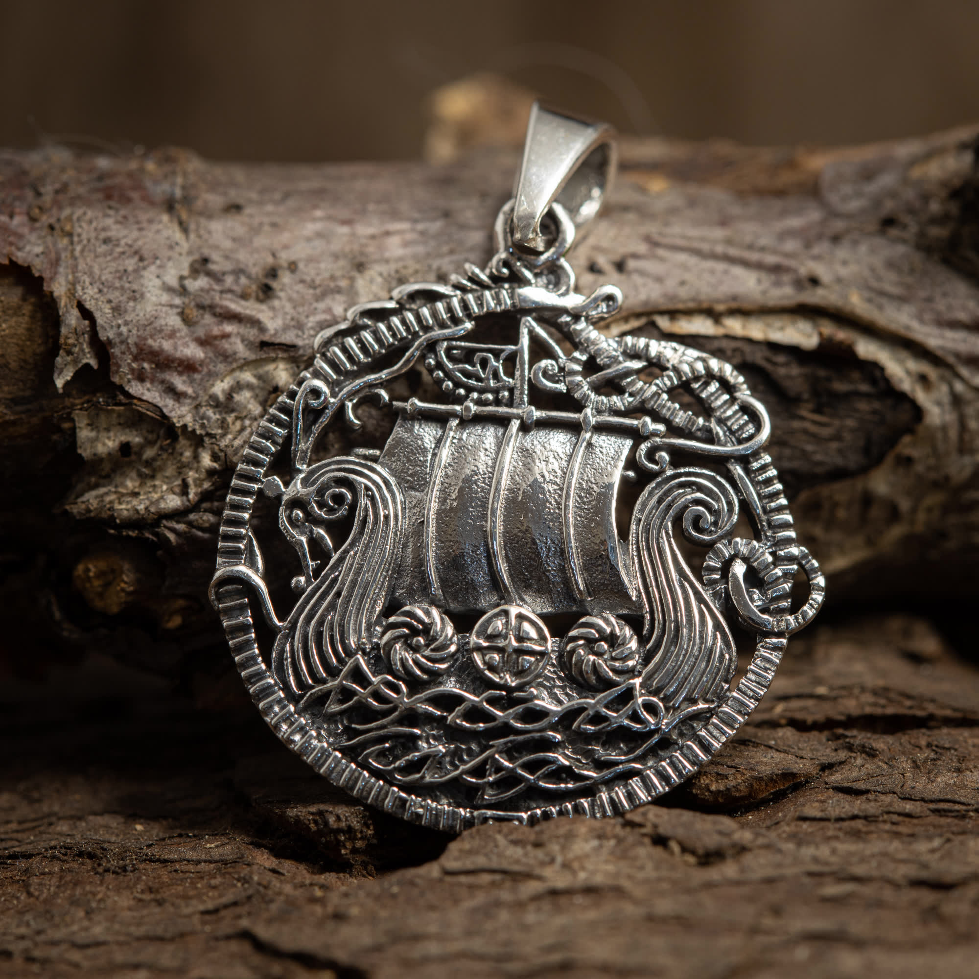 Et nærbillede af Vikingeskib Vedhæng Freya 925s Sterling Sølv, et rundt sølvvedhæng, der viser et indviklet vikingelangskibsdesign med detaljerede sejl, dragehovedbuer og omgivende nautiske motiver. Dette fantastiske smykke af sterlingsølv er vist mod et tekstureret stykke bark.