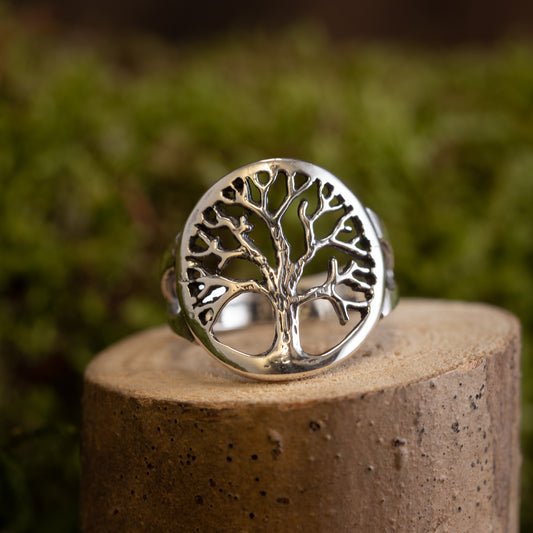 Livets Træ Yggdrasil Fingerring 925s Sterling Sølv viser et indviklet trædesign med bare grene indkapslet i en cirkulær ramme. Dette detaljerede stykke er elegant vist på et lille stykke træ mod en sløret grøn baggrund.