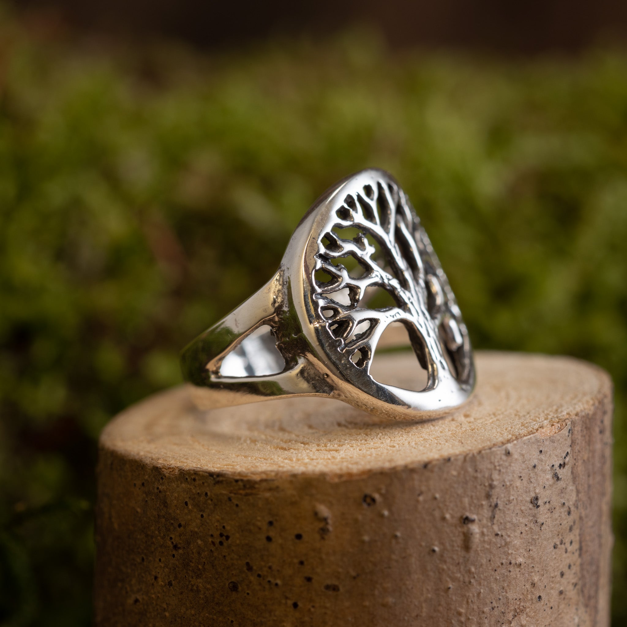 En Livets Træ Yggdrasil Fingerring 925s Sterling Sølv, med et detaljeret træmotiv, vises på en cylindrisk stensokkel. Baggrunden er sløret med grønne nuancer, hvilket tyder på løv eller udendørs landskaber.