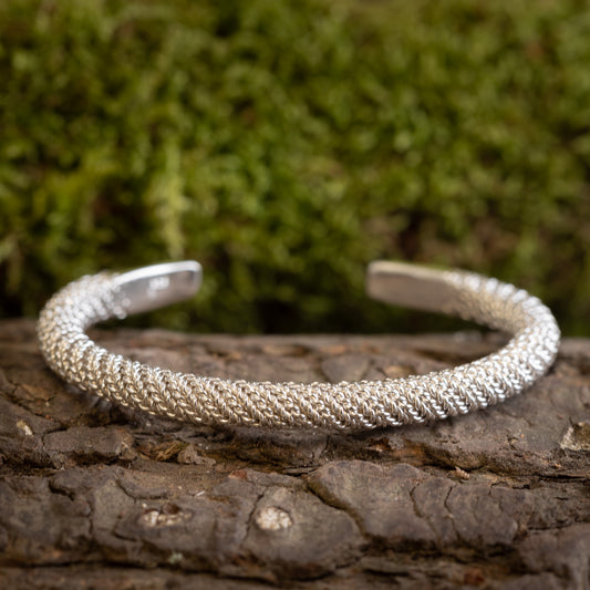 Et Armbånd Bangle Empor 925s Sterling Sølv hviler på et stykke bark, med grønt mos sløret i baggrunden. Armbåndet har et indviklet, vævet design.