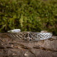 Indlæs billede i Gallery viewer, Et Armbånd Armbånd Mjølnir i 925'erne Sterling Sølv, med indviklede keltiske knudedesigns og et centralt Thors hammersymbol, vises på en træoverflade med en sløret grøn, mosbeklædt baggrund. Det detaljerede håndværk og mønstret er tydeligt synligt.
