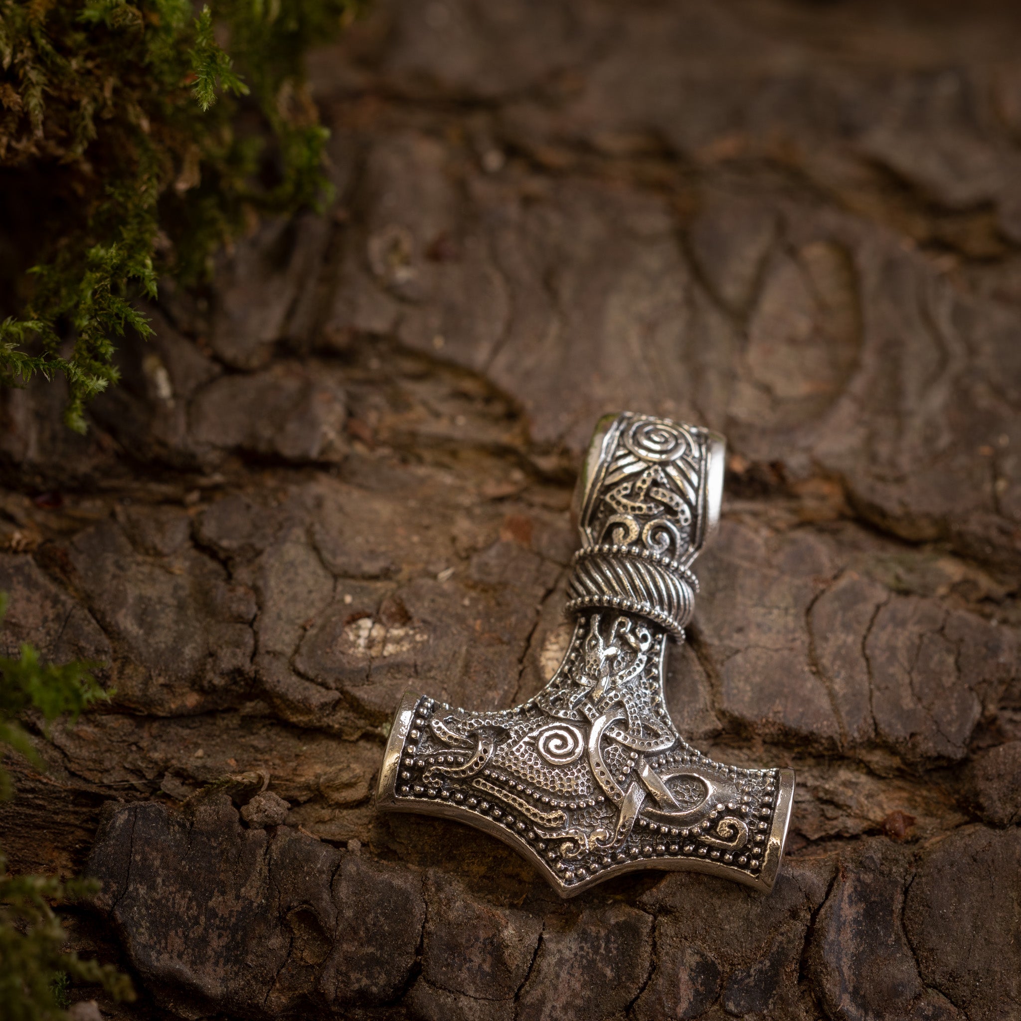 En detaljeret, udsmykket Thors Hammer Vedhæng 925s Sterling sølv Jörmungandr, med indviklede indgraverede mønstre, hviler på et stykke ru træbark. Små totter af grønt mos kan ses i øverste venstre hjørne af billedet.