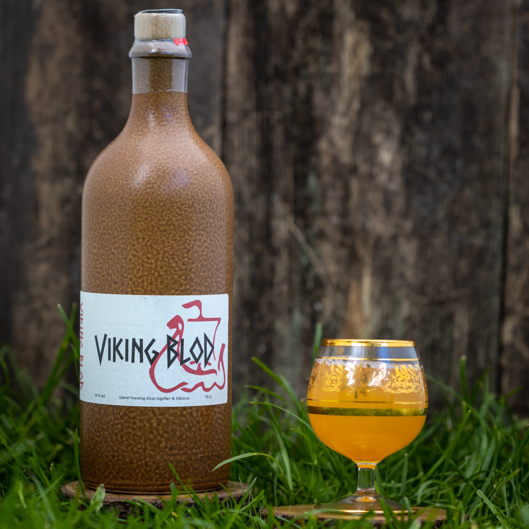 En stor brun flaske mærket "Dansk Mjød Viking Blod (70cl)" med hvid og rød etiket står på græs. Ved siden af står et dekorativt glas fyldt med gylden Dansk Mjød. Et træhegn er sløret i baggrunden, hvilket fremkalder den perfekte velkomstdrink til enhver sammenkomst.