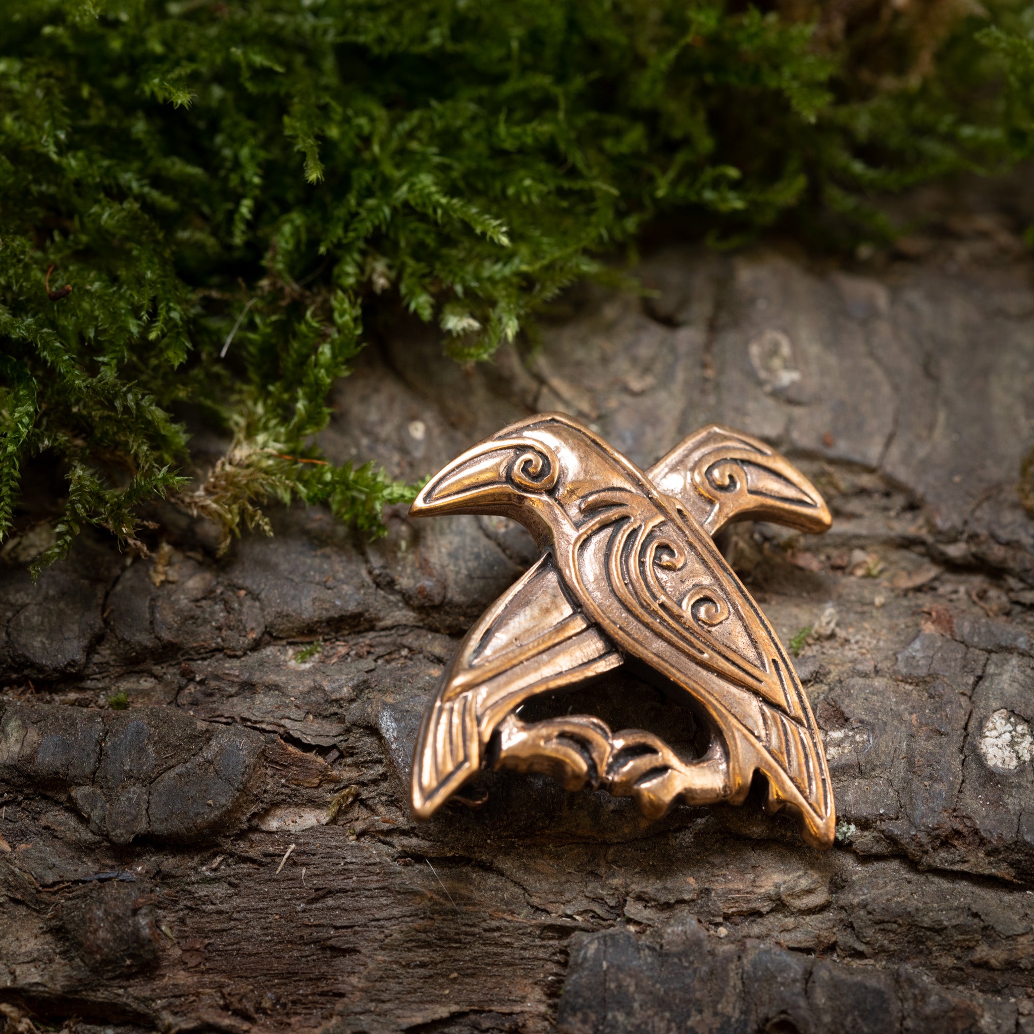 Broche Hugin og Munin Bronze, der er indviklet detaljeret for at fremhæve fjer og former for Odins ravne, hviler på tekstureret træbark med en plet af grønt mos i baggrunden.