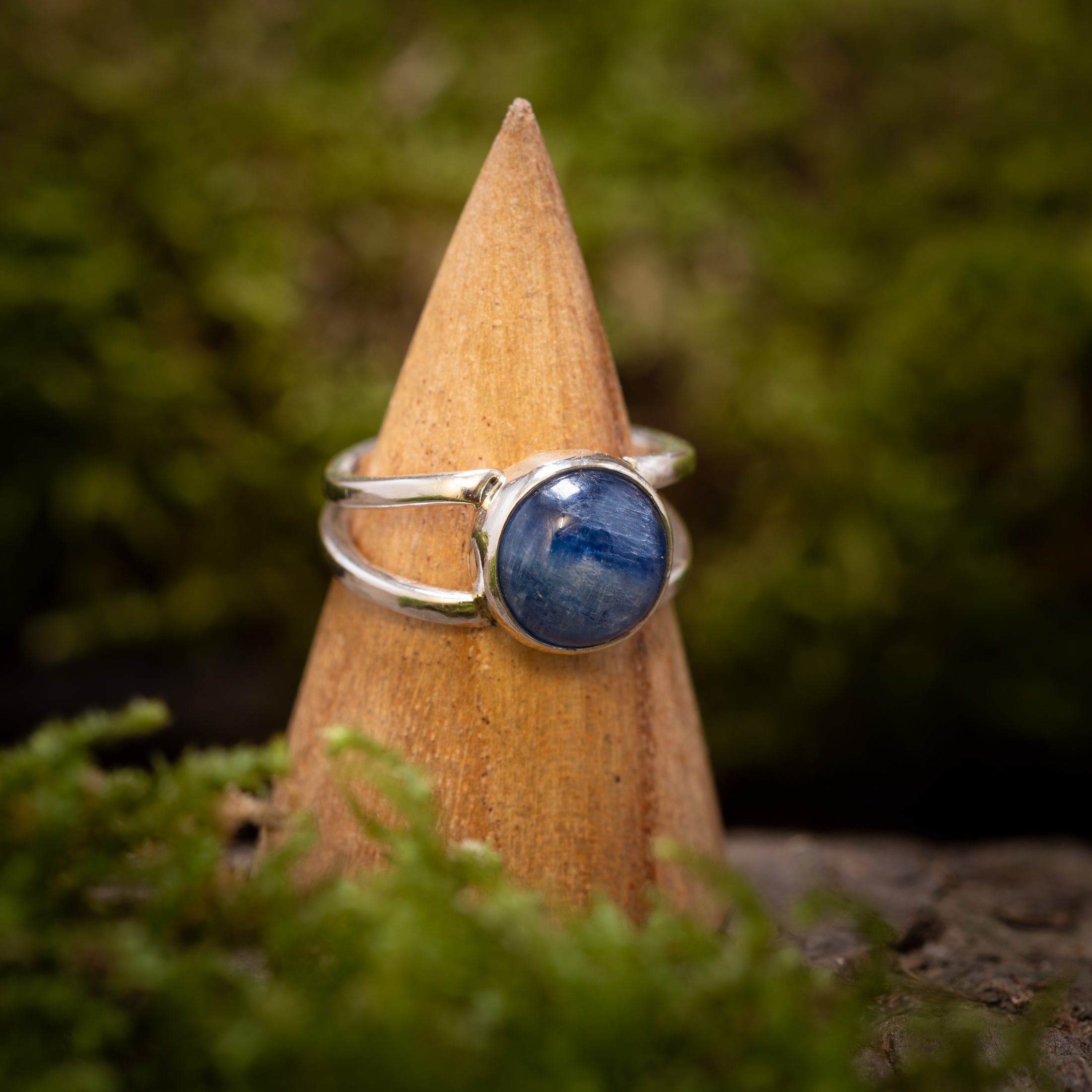 "Fingerring - Eryn Design - Kyanit" er elegant vist på en trækegle, sat mod en sløret grøn baggrund af mos og løv. Dette stykke håndlavede smykker har en fantastisk Kyanit-sten og viser dens 925s Sterling sølv-bånd, som står i smuk kontrast til den levende blå ædelsten og dens naturlige omgivelser.