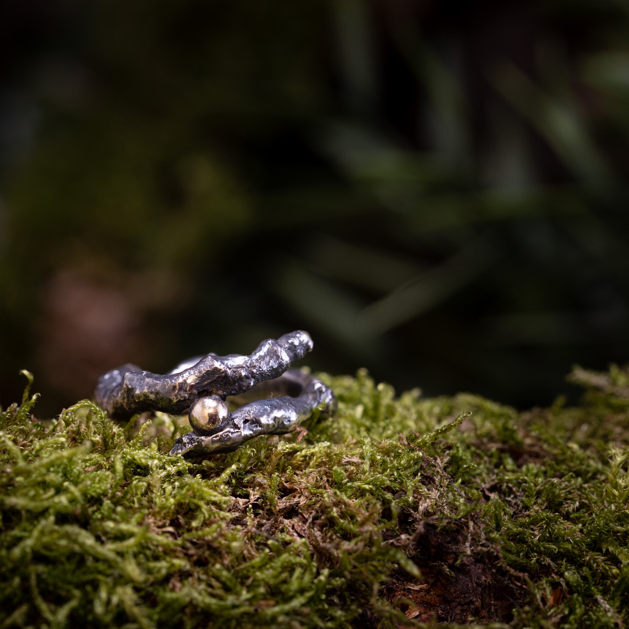 Et nærbillede af den udsøgt udformede Fingerring - Eryn Design - Treasure, lavet af 925'er Sterling Sølv og med en delikat guldkugle, hviler på levende grønt mos. Den let slørede baggrund fremhæver ringen og mosens naturlige tekstur.